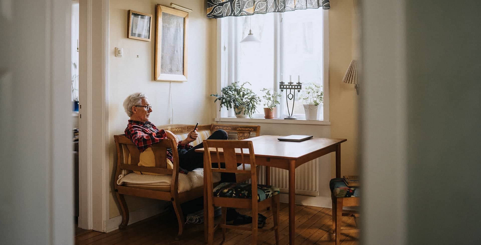 En bild på en äldre man som sitter ensam med sin telefon vid ett köksbord.