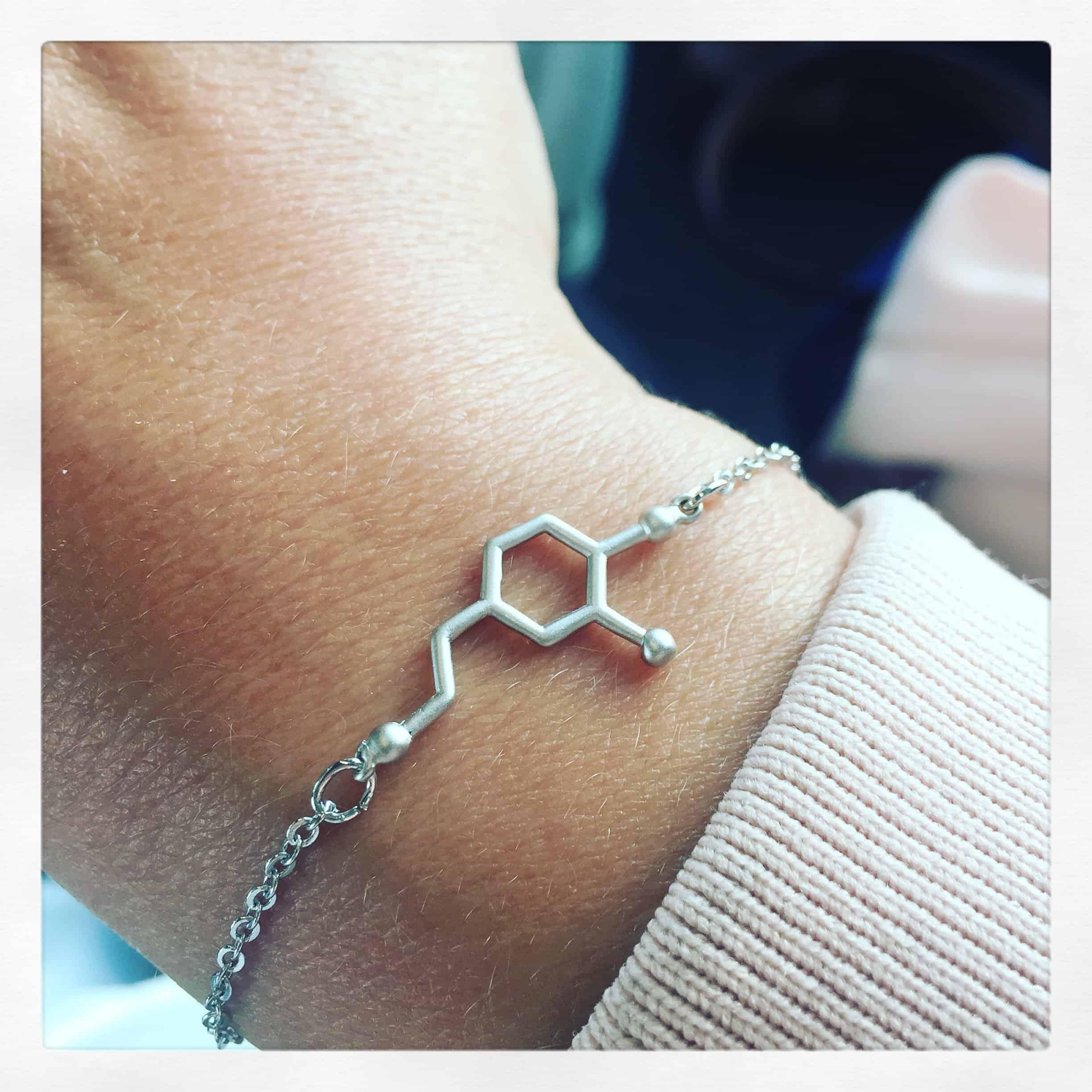 Närbild på en handled som bär ett silvrigt armband med en symbol som föreställer en dopaminmolekyl.