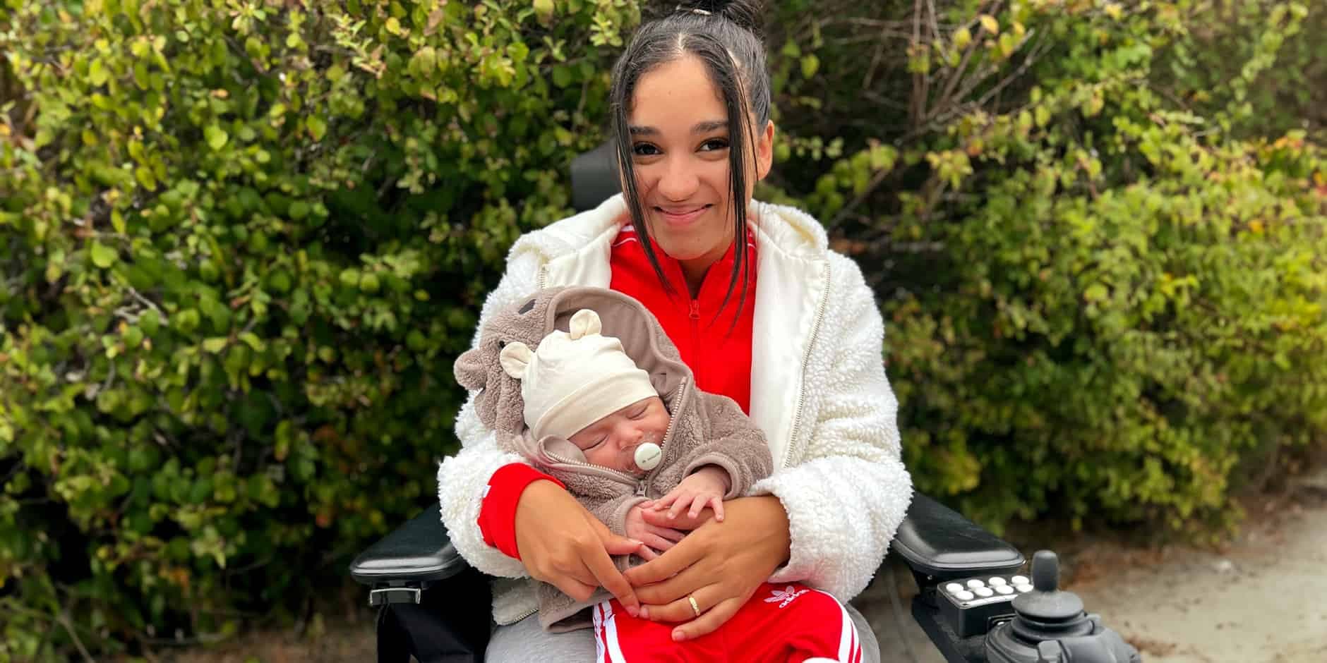 Carolina sitter framför ett buskage i sin rullstol med sin son Matheus i famnen