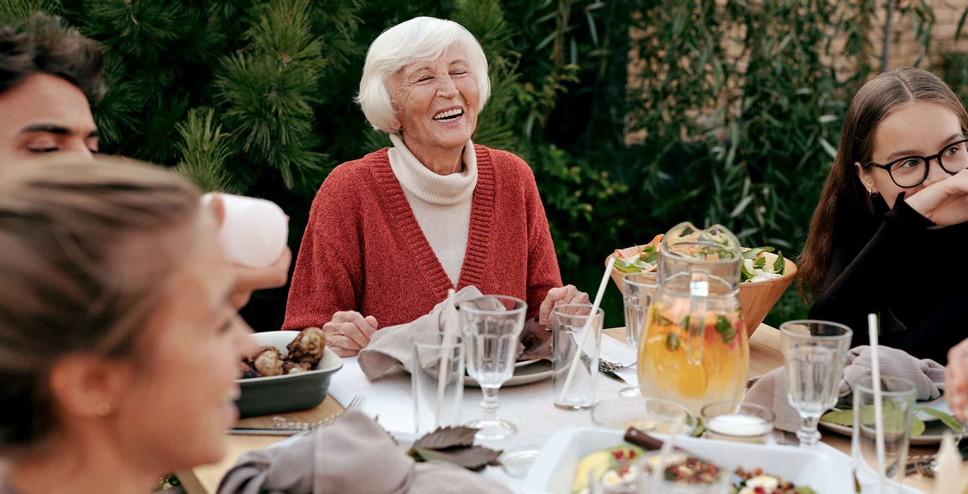 Fokus på äldre dam som sitter vid ett bord utomhus tillsammans med bekanta och ler.