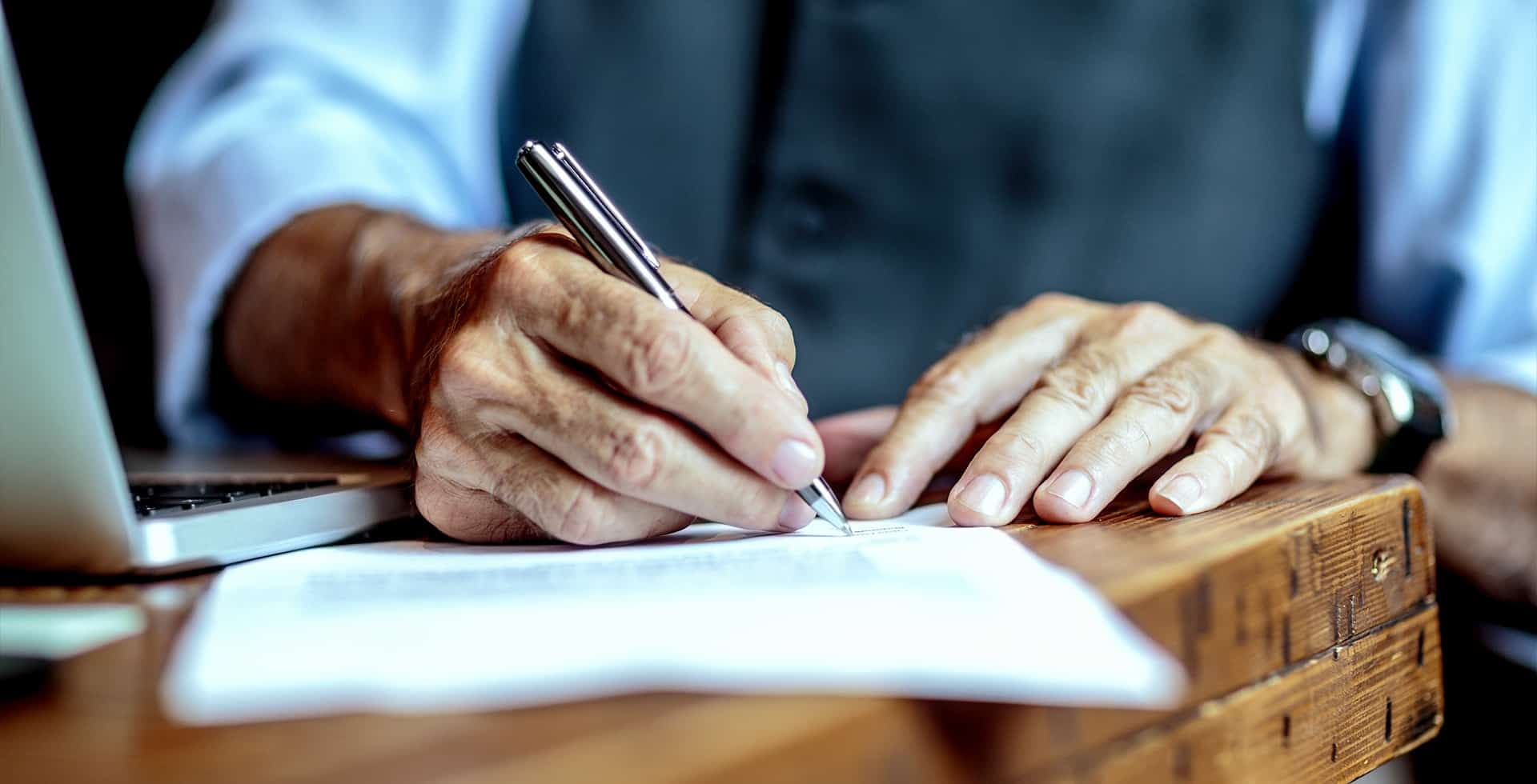 Foto på en persons händer som håller i en penna medan den antecknar på ett dokument som ligger på ett bord.