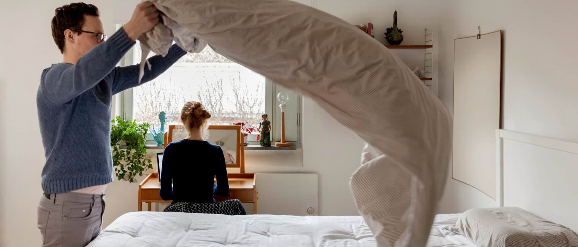 En person bäddar sängen genom att lägga på ett täcke, medan en annan person sitter vid ett skrivbord och arbetar med en laptop. Rummet är ljust och inrett med växter och konstnärsmaterial.
