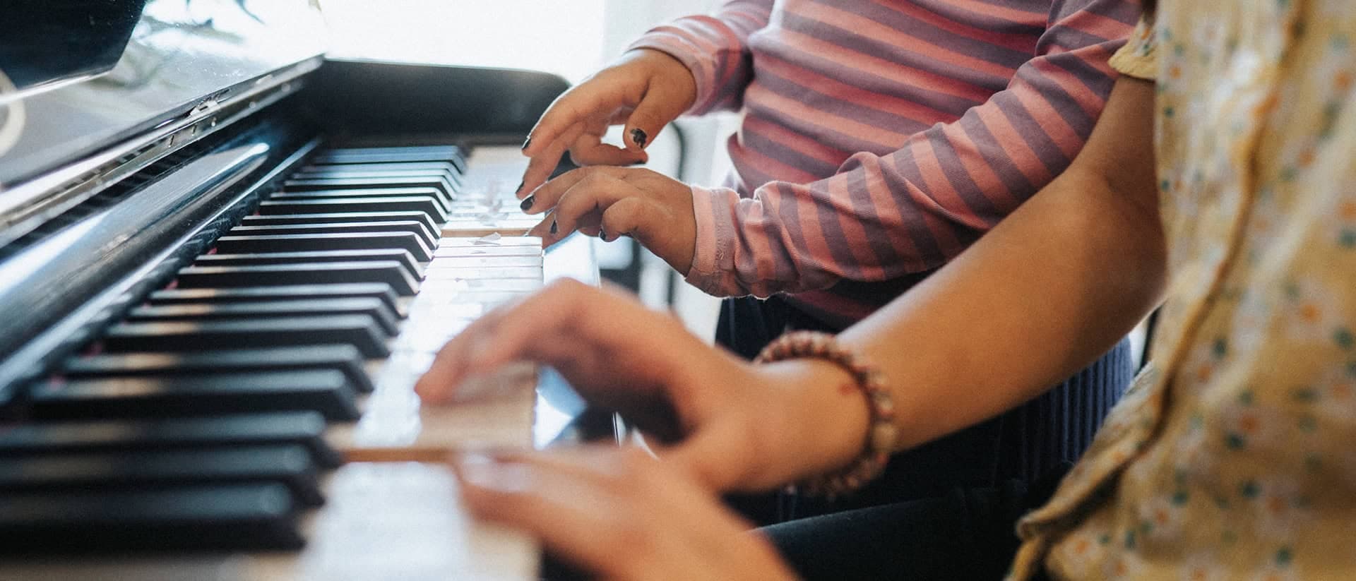 Två personer spelar piano tillsammans, deras händer rör sig över tangenterna. Den ena personen bär en randig tröja, medan den andra har en mönstrad topp. Ljuset från ett fönster skapar en varm och kreativ atmosfär.