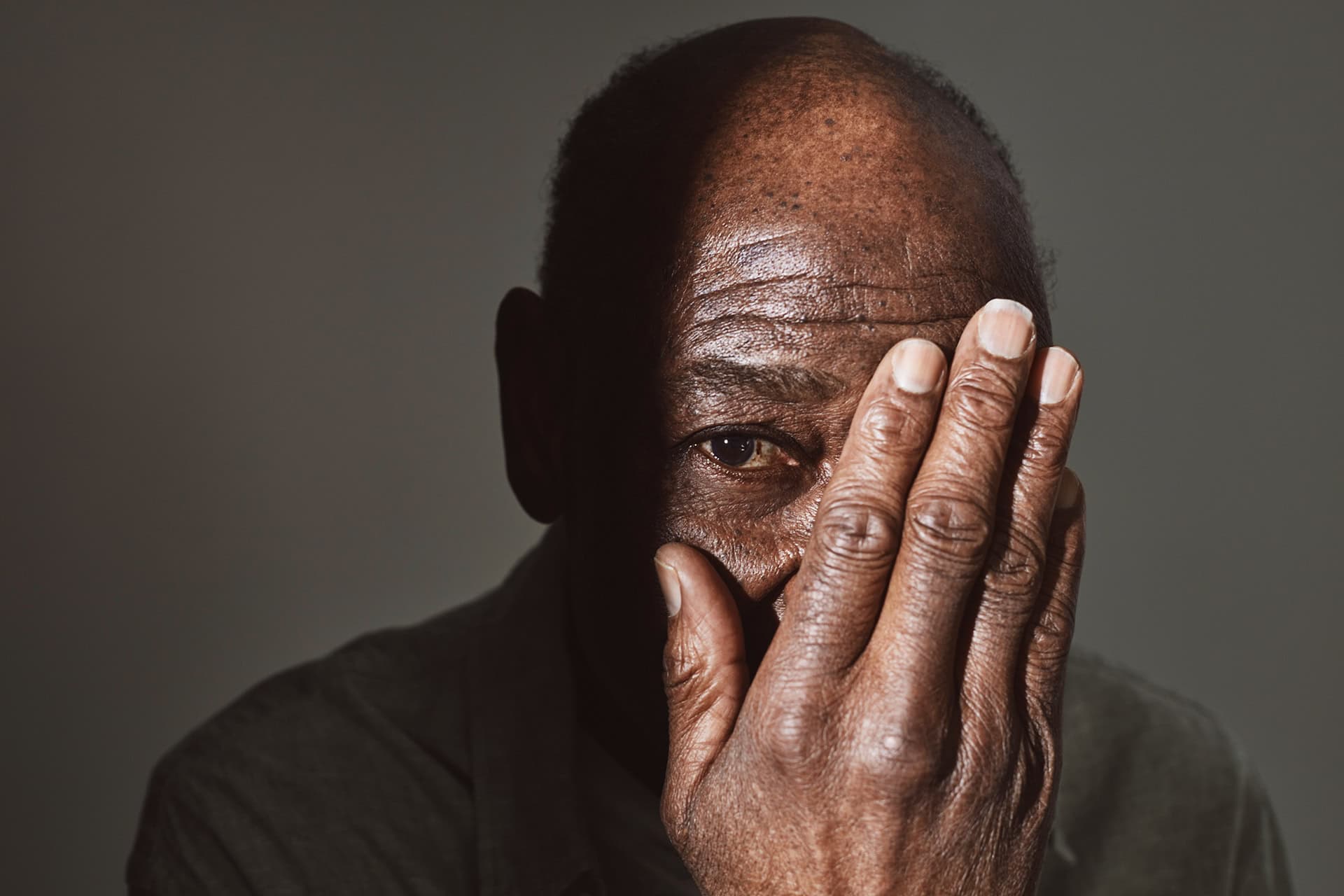 En äldre man täcker delvis sitt ansikte med handen, medan han ser allvarsamt in i kameran. Bilden har en dämpad och eftertänksam känsla.