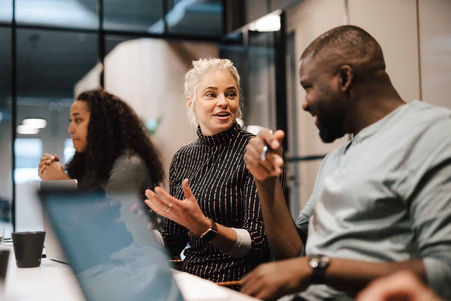 Tre personer sitter runt ett bord i en samtalssituation. En kvinna med ljus hår knuten i en knut pratar engagerat med en man mittemot, medan en tredje person lyssnar i bakgrunden. Miljön är modern och professionell.