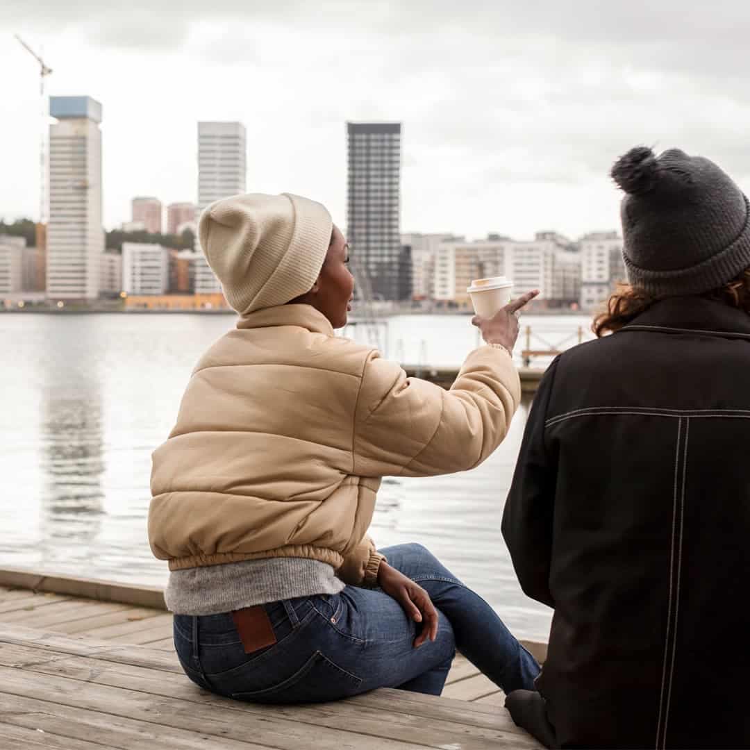 Två personer sitter på en brygga vid vattnet i en urban miljö, iförda vinterkläder och mössor. Den ena personen gestikulerar med en kaffekopp i handen, medan de samtalar och blickar ut över stadens höga byggnader.