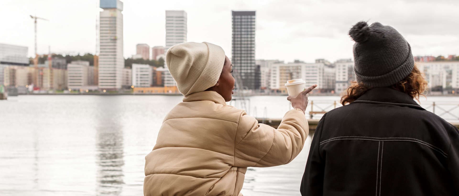 Två personer sitter på en brygga vid vattnet i en urban miljö, iförda vinterkläder och mössor. Den ena personen gestikulerar med en kaffekopp i handen, medan de samtalar och blickar ut över stadens höga byggnader.