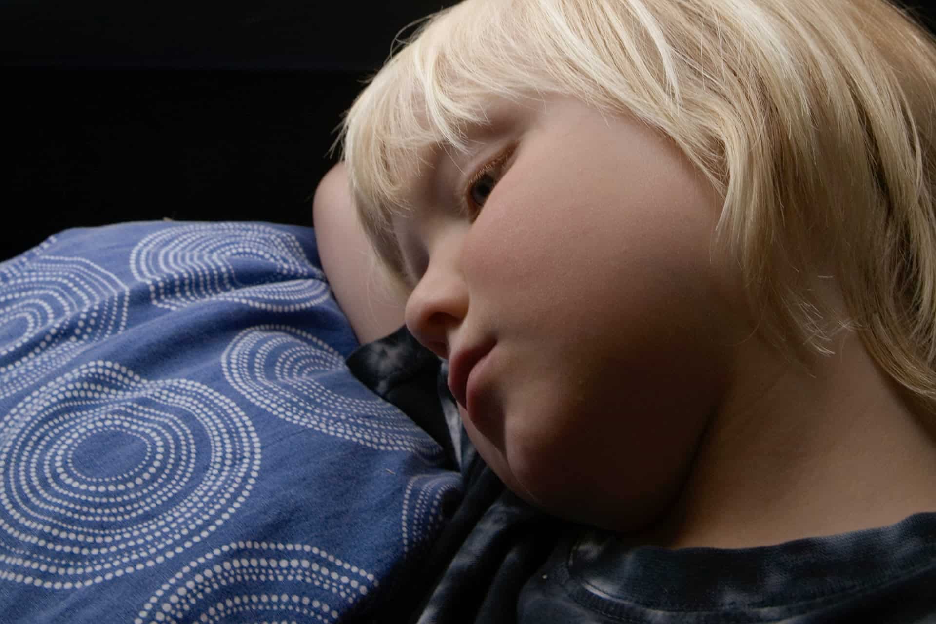 Ett barn med blont hår ligger på en kudde med blått mönstrat örngott och ser tankfullt ut. Miljön är dämpad och lugn.