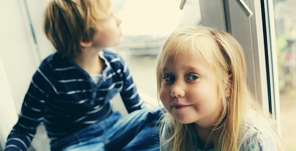 En flicka i sjuårsåldern tittar in i kameran. I bakgrunden sitter hennes lillebror och ser ut genom fönstret.
