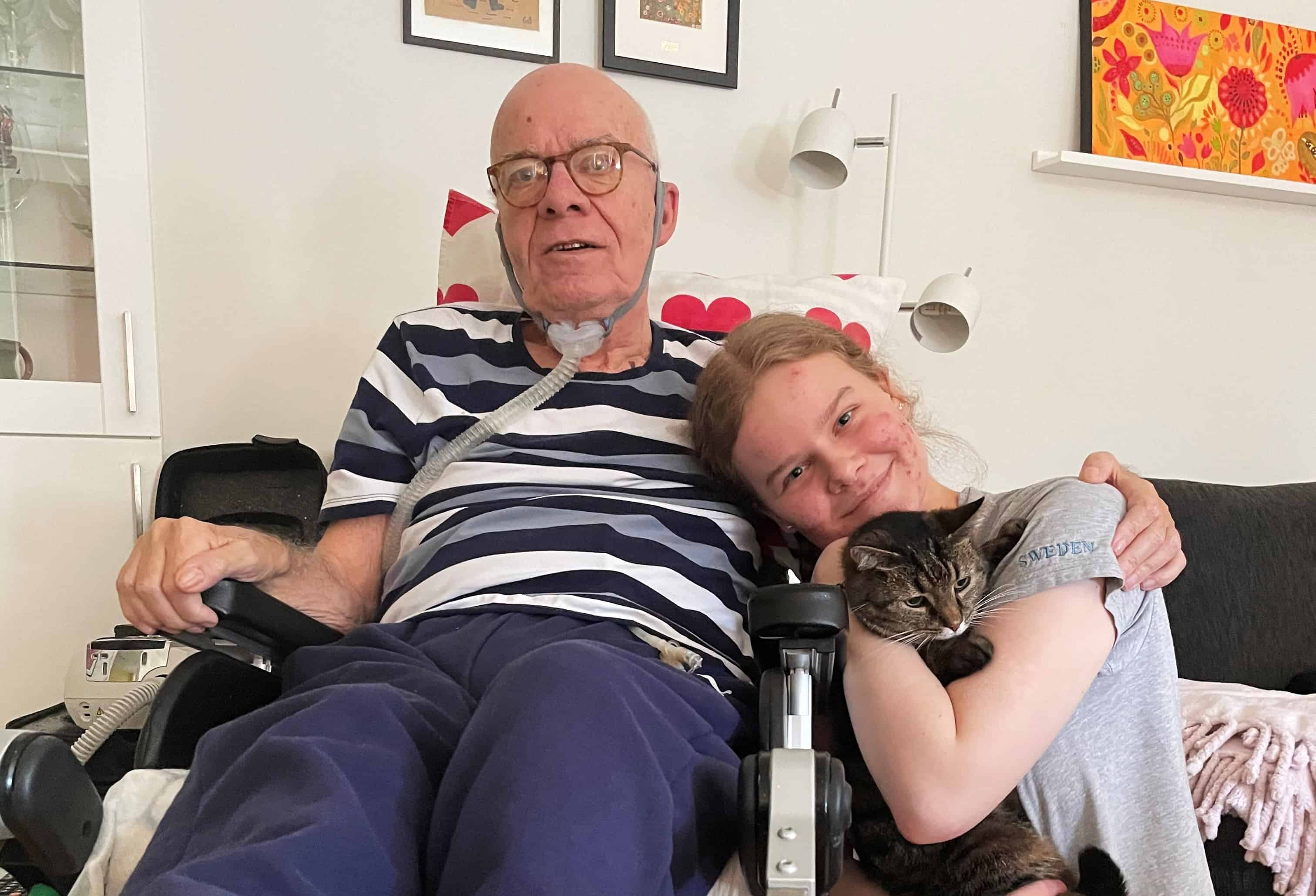 En äldre man i rullstol. Intill honom sitter hans barnbarn med en katt i famnen. Hon lutar sig mot morfar.